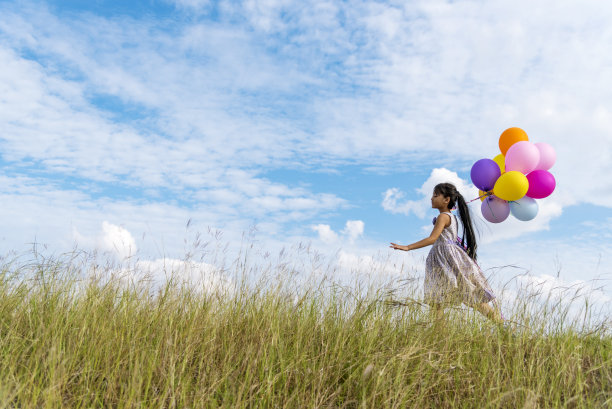 活泼可爱的女孩拿着气球在绿色的草地上奔跑，白云和蓝天上洋溢着幸福。手握充满活力的气球在生日派对上玩快乐时光夏天在户外的阳光图片下载