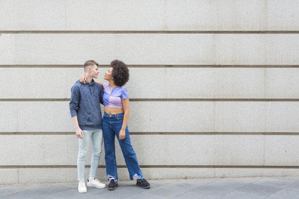 一名年轻的白人男子和一名黑人女子倚靠在一堵灰色的墙上互相凝视着对方的眼睛图片下载