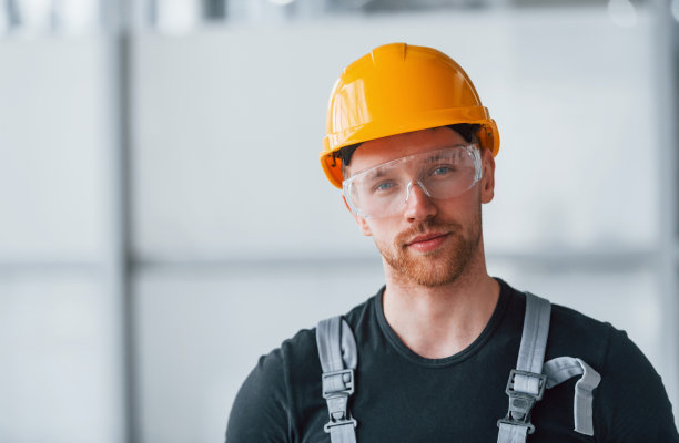 白天站在现代大办公室的室内，穿着灰色制服，戴着防护眼镜和橙色安全帽的男人的肖像图片下载