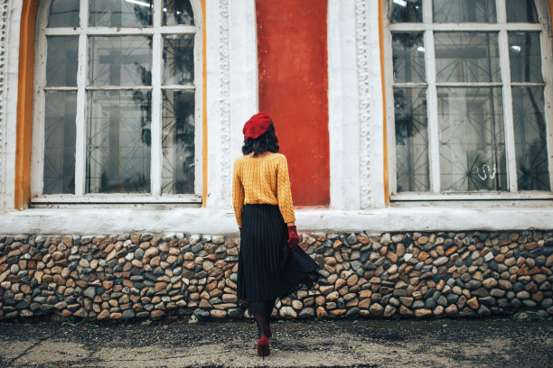 一个穿着黄色毛衣，戴着红色帽子的女人站在一个有大窗户的建筑物的背景下图片下载
