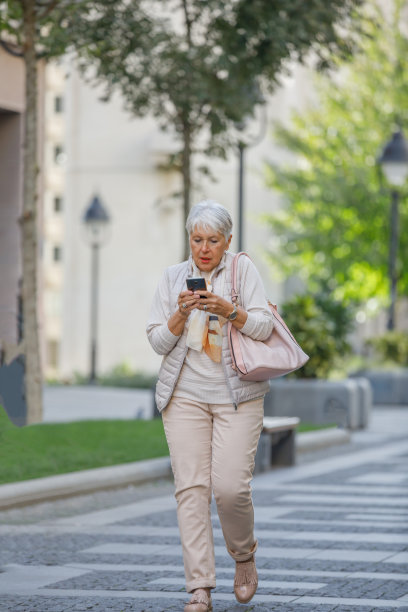 一位老年妇女走在城市的街道上，用手机输入信息。图片下载