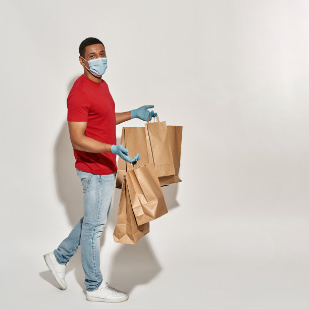 在浅灰色的背景下，一名快乐的快递员戴着防护面具和手套，拿着许多棕色的牛皮纸袋准备外卖图片下载