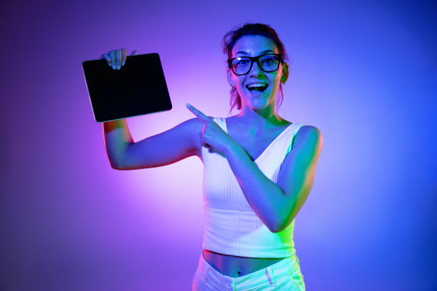 拿着平板电脑，疯狂兴奋。在霓虹灯下的黑色画室背景上的白人年轻女子肖像。穿着白衬衫的漂亮男模特。图片下载