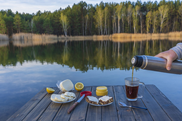 早上在湖边和森林里，把热茶从保温瓶里倒进玻璃杯里。早餐在木桌上图片下载