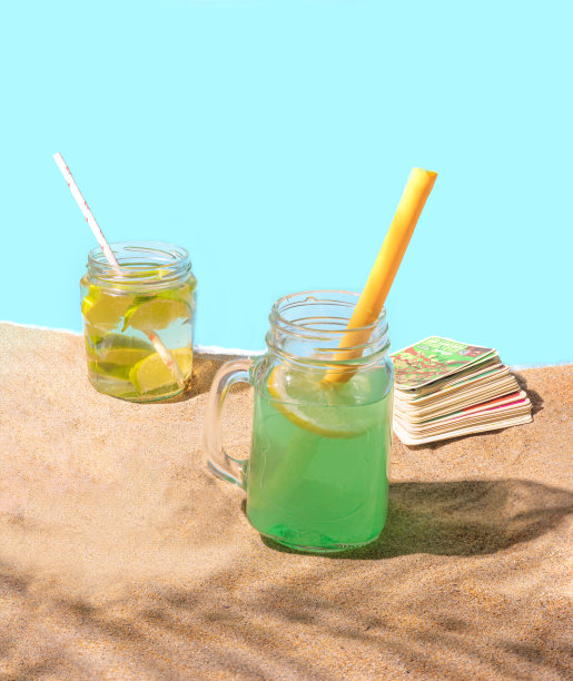 两个装着鸡尾酒饮料和纸的罐子和可重复使用的硅胶吸管放在沙滩上，棕榈叶的阴影，背面是扑克牌。环保理念，环保安全度假图片下载