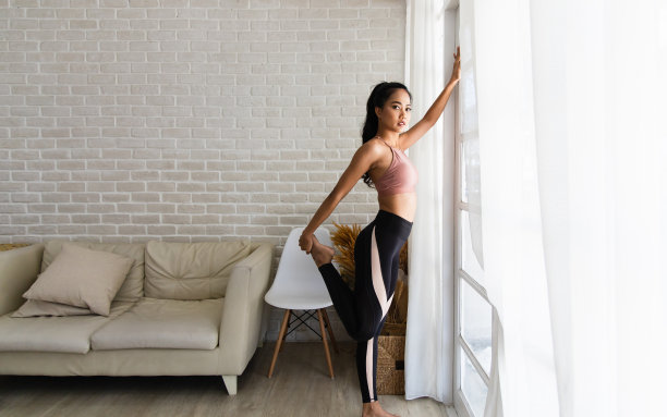 一个美丽的亚洲女人在家健身而不是去健身馆，她在白色的房间里热身伸展双腿。她穿着运动装。锻炼的概念好身材图片下载