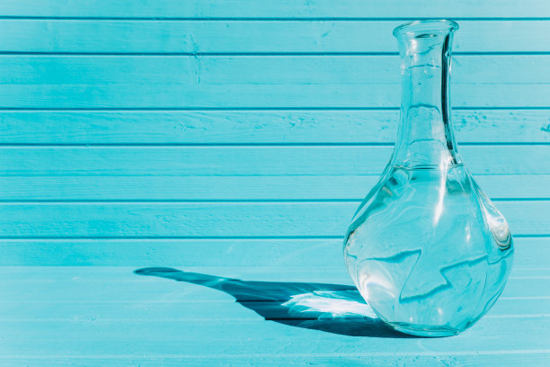 极简主义风格的静物-一个玻璃花瓶与水在绿松石木制背景。太阳照射在一个反射光线的透明容器上图片下载