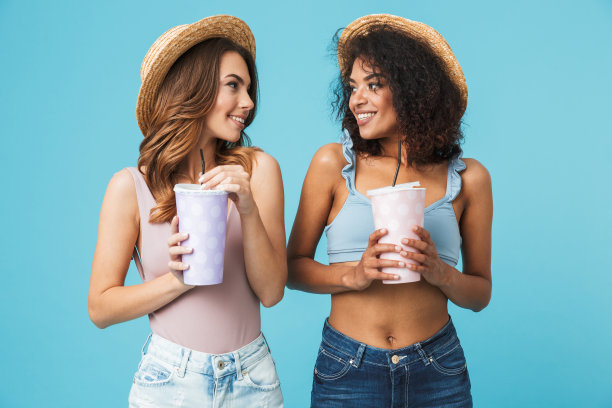 戴着草帽的两名20多岁的年轻貌美的女性，在蓝色的背景下，拿着装苏打水或冷饮的纸杯，互相看着对方图片下载