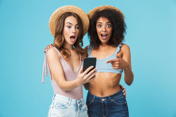 两个多民族夏日女孩的度假照片，在蓝色背景下，张着嘴，手指着智能手机屏幕自拍，表达兴奋图片下载