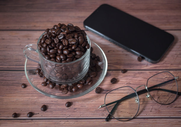 智能手机和透明的玻璃杯，装满烘焙香气的咖啡豆和光过滤和眼睛保护眼镜。工作业务的概念。图片下载