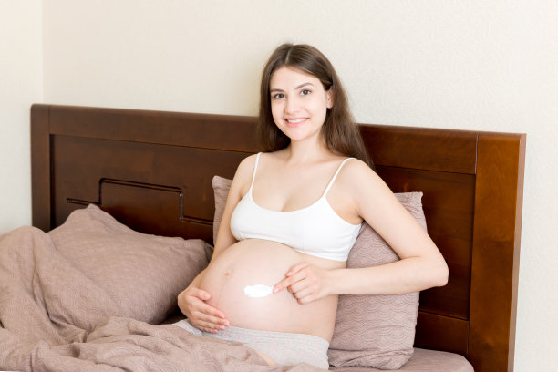 一个怀孕的女孩坐在家里的床上，把抗妊娠纹霜涂在她的肚子上。怀孕、母性、准备和期望的概念图片下载