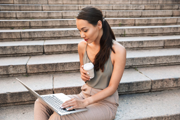照片中，20多岁的漂亮年轻女子穿着休闲的夏装，坐在城市的楼梯上，一边使用银色笔记本电脑一边喝外卖咖啡图片下载