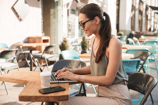 照片中，20多岁的白人女性戴着黑色太阳镜坐在城市的咖啡馆或户外餐厅里，使用银色笔记本电脑图片下载