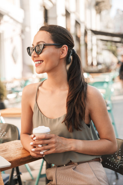 照片上，20多岁的年轻女性戴着黑色太阳镜，穿着夏装，微笑着坐在城市的咖啡馆或户外餐厅，用纸杯喝咖啡图片下载