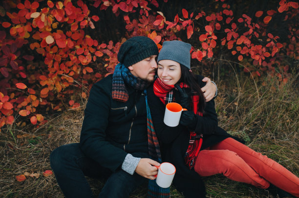 一对相爱的年轻夫妇穿着大衣，戴着帽子，围着围巾，坐在红色的秋天灌木丛旁的草地上喝着咖啡。在大自然中休息和放松的概念。图片下载
