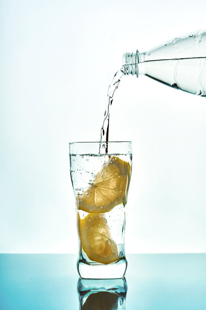 将蓝色背景的塑料瓶中的柠檬水倒入玻璃杯中。新鲜的水杯与泡沫蓝色背景。图片下载