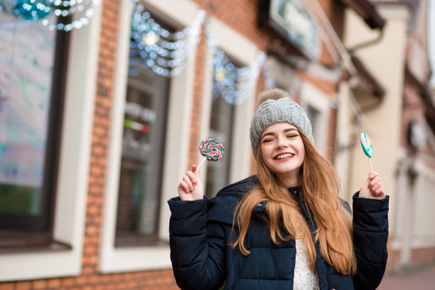 红色头发的女人戴着灰色针织帽，拿着圣诞糖果靠近商店的橱窗图片下载