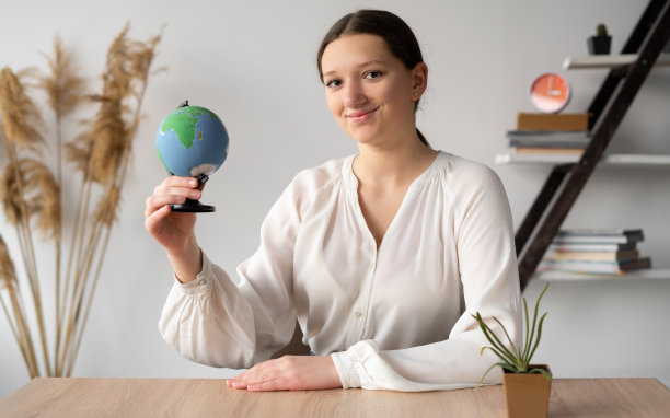 办公室里微笑着的女孩手里拿着地球仪组织了一次旅行。组织者通过网络摄像头显示旅行地点。旅游概念。图片下载