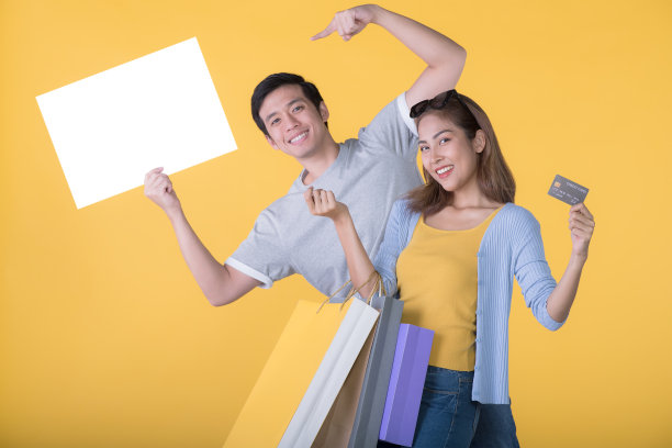 一对亚洲夫妇手持空白广告牌，上面有信用卡和购物袋，背景是黄色图片下载