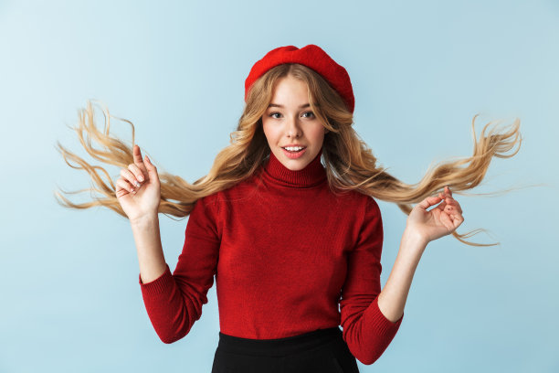 优雅的金发女人20多岁的照片戴着红色贝雷帽，挥舞着她的头发，而站着，孤立在工作室的蓝色背景图片下载