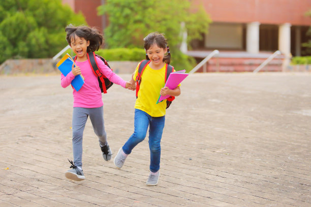 2019冠状病毒病流行后的返校生活。两个可爱的小女孩一边走路一边说话，手里拿着书，肩上扛着书包去上学。图片下载