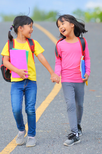 2019冠状病毒病流行后的返校生活。两个可爱的小女孩一边走路一边说话，手里拿着书，肩上扛着书包去上学。图片下载