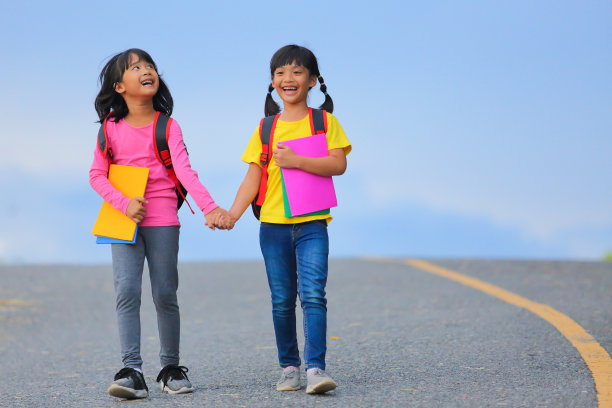 2019冠状病毒病流行后的返校生活。两个可爱的小女孩一边走路一边说话，手里拿着书，肩上扛着书包去上学。图片下载