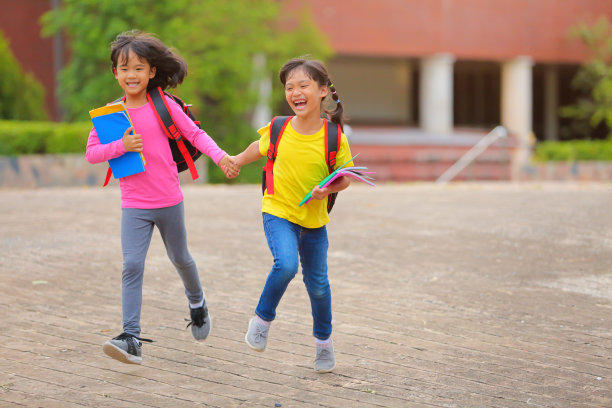 2019冠状病毒病流行后的返校生活。两个可爱的小女孩一边走路一边说话，手里拿着书，肩上扛着书包去上学。图片下载