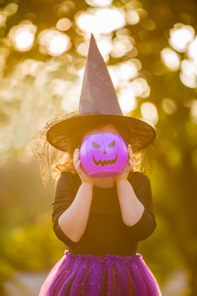 万圣节快乐。在秋天的森林里，一个戴着女巫帽，带着一桶紫色南瓜糖果的微笑女孩的肖像。欢乐的孩子们穿着狂欢节的服装在大自然中图片下载