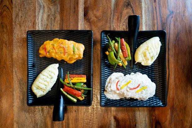 平面照片显示了两个美观的黑色方形带把手的盘子放在木桌上，里面装满了白酱意大利面和红酱土豆泥和辣椒蔬菜沙拉图片下载