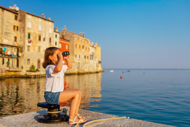 一个有趣的小女孩在阳光明媚的夏天用老式望远镜看东西。罗温镇的背景。图片下载