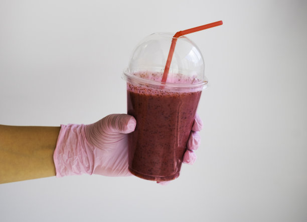 手在一个粉红色的橡胶手套拿着一杯浆果冰沙。大流行时期的医疗保健概念图片下载