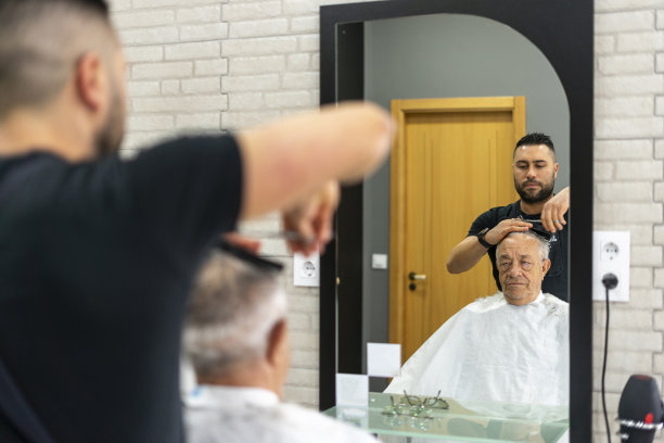 一个年轻的理发师正在用从理发店的镜子里映出的剪刀给一个老人理发。图片下载