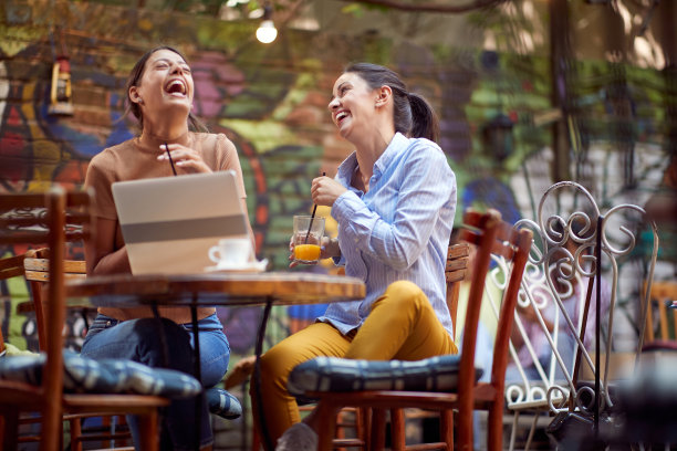 两个年轻的成年女性朋友坐在开着门的咖啡馆里，大声笑了起来。图片下载