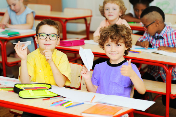 两个学前班的小男孩坐在教室的课桌上，手里拿着纸飞机，微笑着看着一群小学同学图片下载