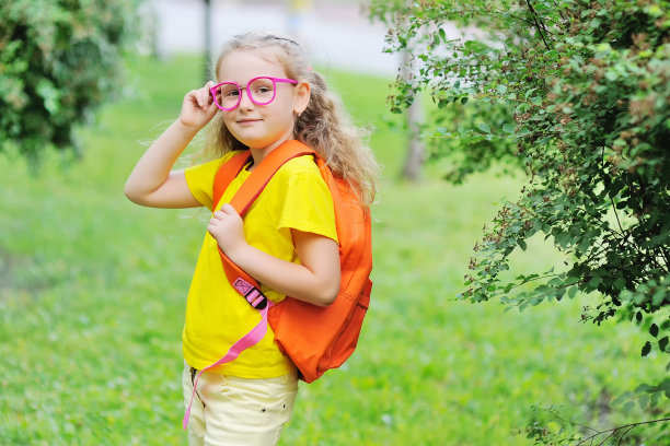 一名身穿黄色t恤、戴粉色眼镜、背着书包的小女生，在公园和绿树的映衬下露出微笑。回到学校，教育，知识日，学习用品图片下载