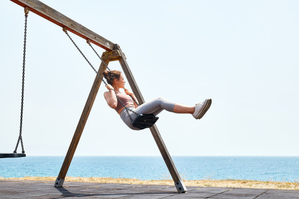 漂亮的年轻女子，穿着运动服，在海边的操场上荡秋千。自由和无忧无虑的情绪。回到童年。图片下载