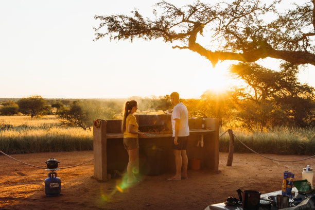 年轻的女人和男人露营与露营车在喀拉哈里沙漠享受日落和准备晚餐在纳米比亚库存照片图片下载