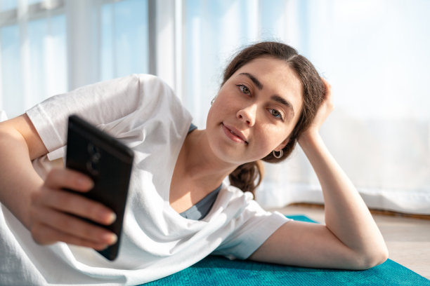 一幅年轻女子微笑着躺在运动垫上使用智能手机的照片。背景中的Windows。通信和网络图片下载