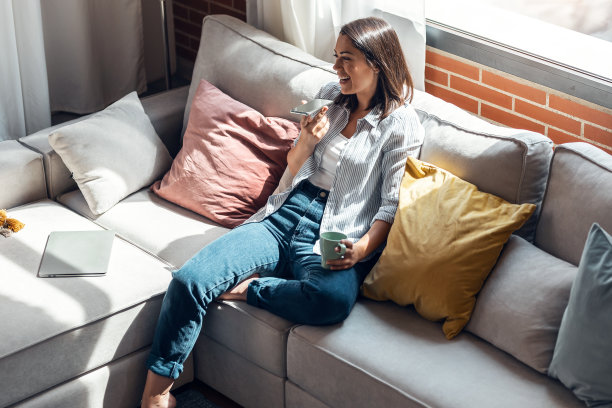 一个年轻漂亮的女人坐在家里的沙发上玩手机。图片下载