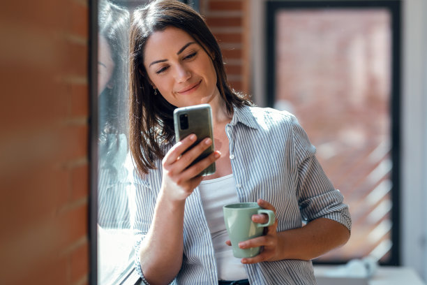 美丽的年轻女子使用她的手机，而站在家里的窗户旁边。图片下载