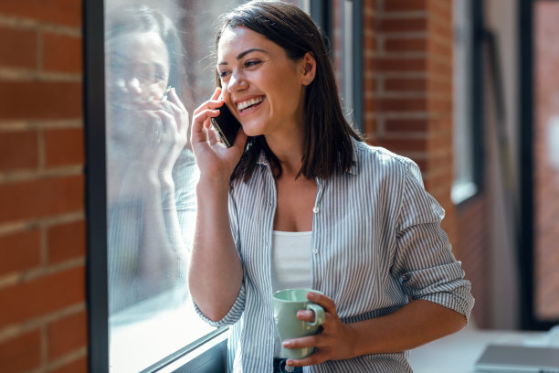 美丽的年轻女子使用她的手机，而站在家里的窗户旁边。图片下载