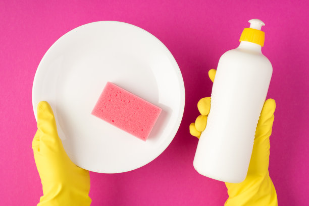 俯视图照片的手在黄色橡胶手套拿着白色洗涤剂瓶没有标签白色清洁板和粉红色洗涤垫孤立的粉红色背景与copyspace图片下载