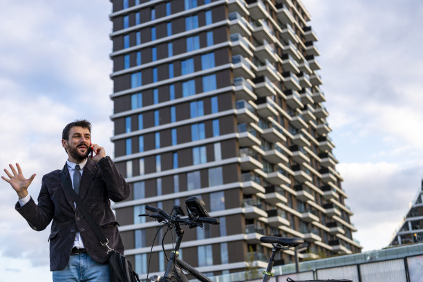 一个年轻时尚的商人正在骑自行车上班，并且正在用他的手机聊天。微笑的男人在街上用智能手机。图片下载