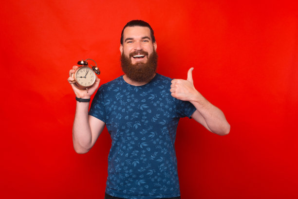 红色背景下，一名身穿蓝色t恤的快乐大胡子男子举着闹钟，拇指朝上图片下载