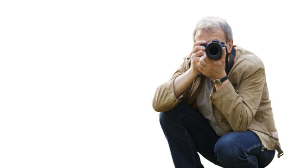 一位中年男子拿着相机蹲着拍照。在前景中，照相机对准观众。一位经验丰富的摄影师，被孤立在白色背景上。本空间图片下载
