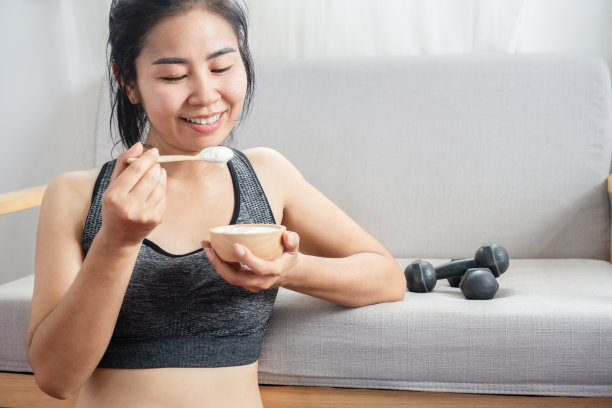 亚洲运动女孩吃酸奶健身后在家锻炼，旁边坐着哑铃图片下载