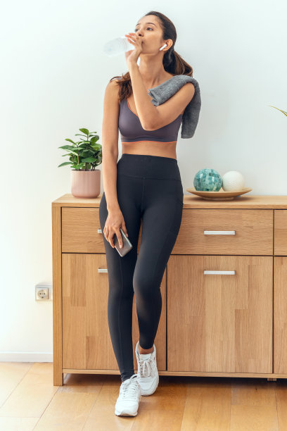 这张照片拍摄的是一位年轻的运动女性在家里的客厅里做运动休息的时候喝水。图片下载