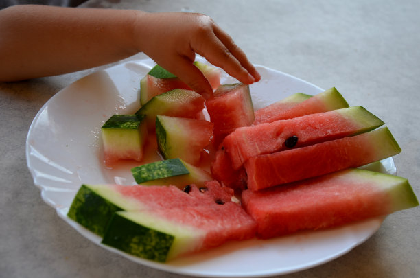 一个小男孩的手伸向一个西瓜。桌上放着切好的西瓜。红色果肉和黑色种子。图片下载