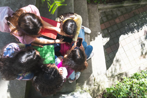 这张照片拍摄的是一群十几岁的学生朋友坐在一个露天看台上，手里拿着手机、背包和笔记本图片下载